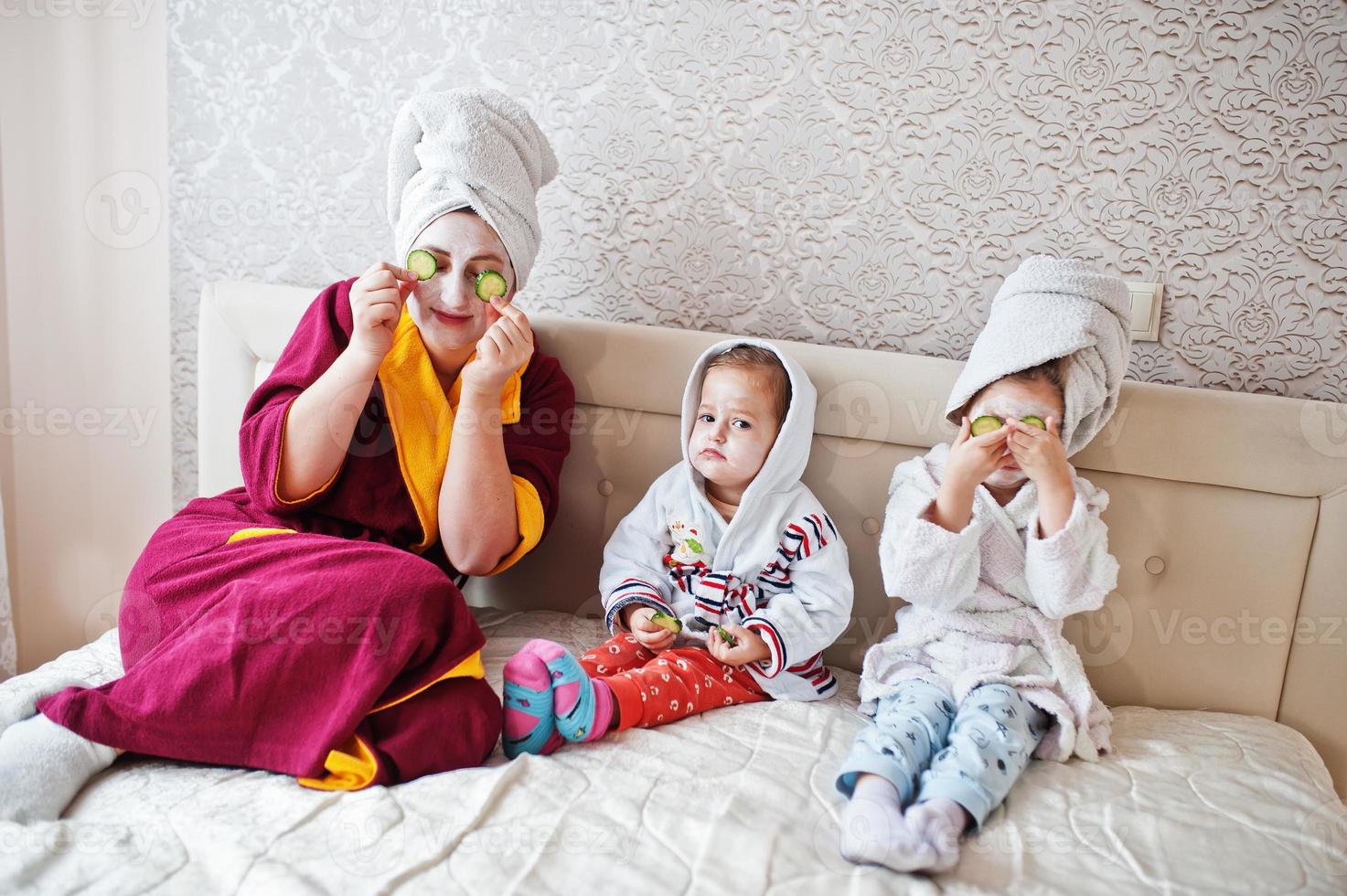 Mother with daughters in a bathrobe with a towel on her head take care of the skin at home, cucumbers on eyes. photo