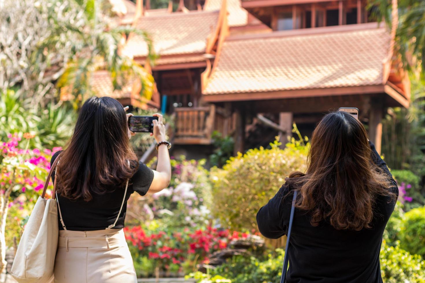 Looking behind the two young Thai women taking pictures with their mobile phones to an old wooden Thai house in a park during the day. photo