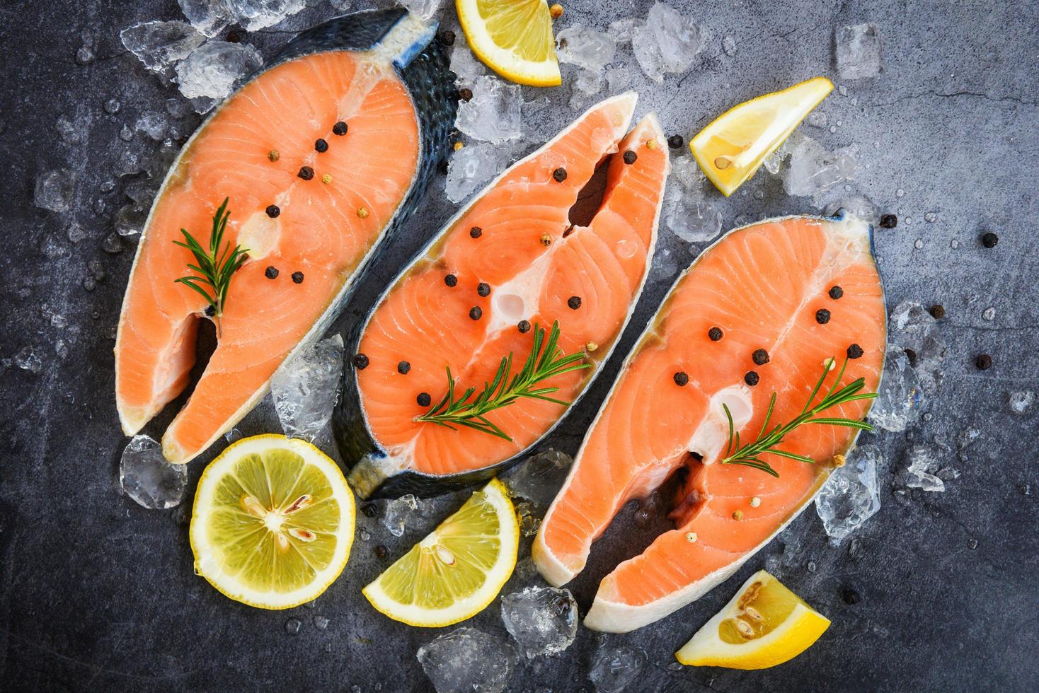 Fresh salmon steak with herbs and spices lemon rosemary on black plate background Raw salmon fish fillet and ice for cooked steak seafood photo