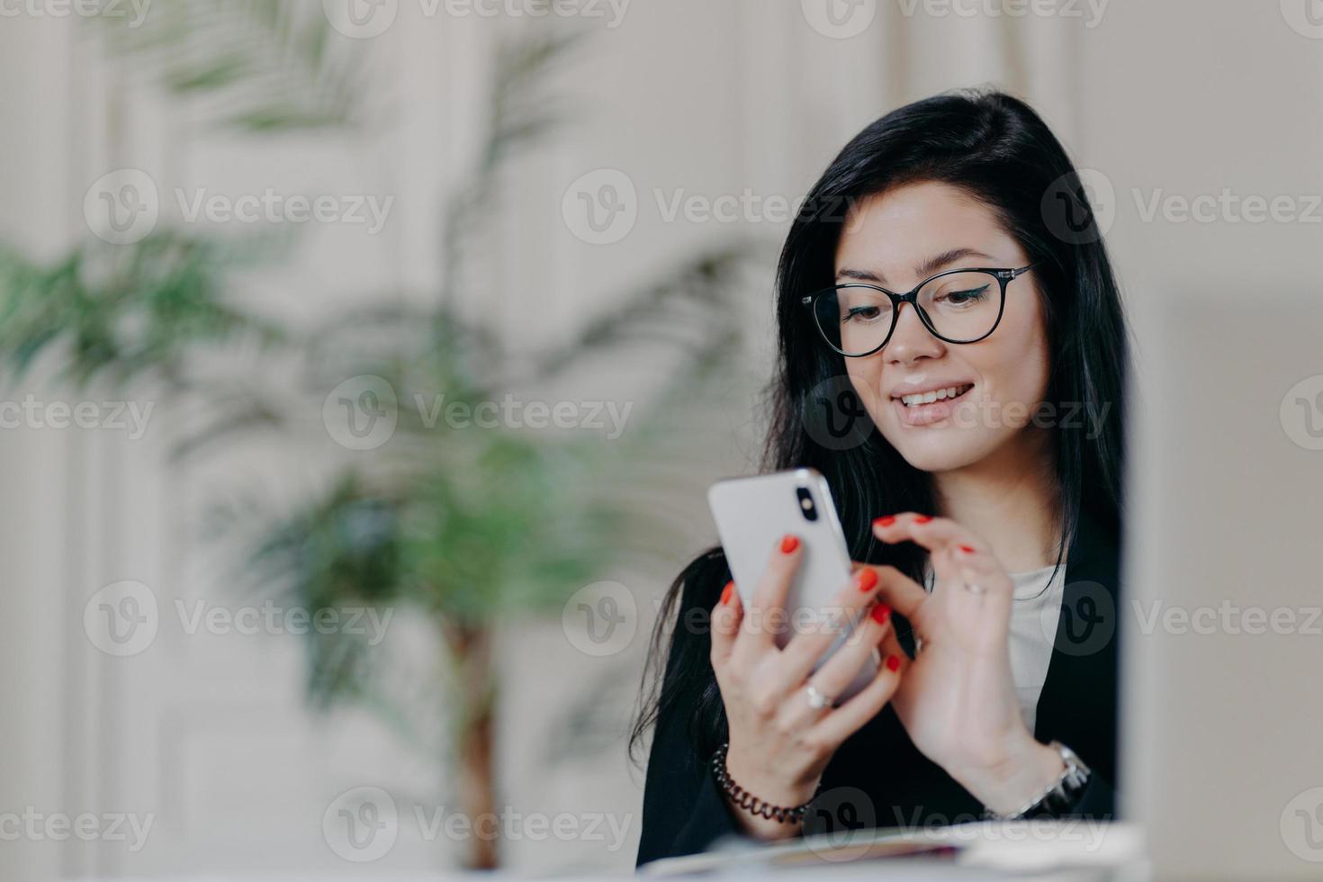 Happy brunette businesswoman uses smartphone, wears spectacles, sends text messages to partner, works on project, poses against cozy interior at workplace. Technology, occupation and work concept photo