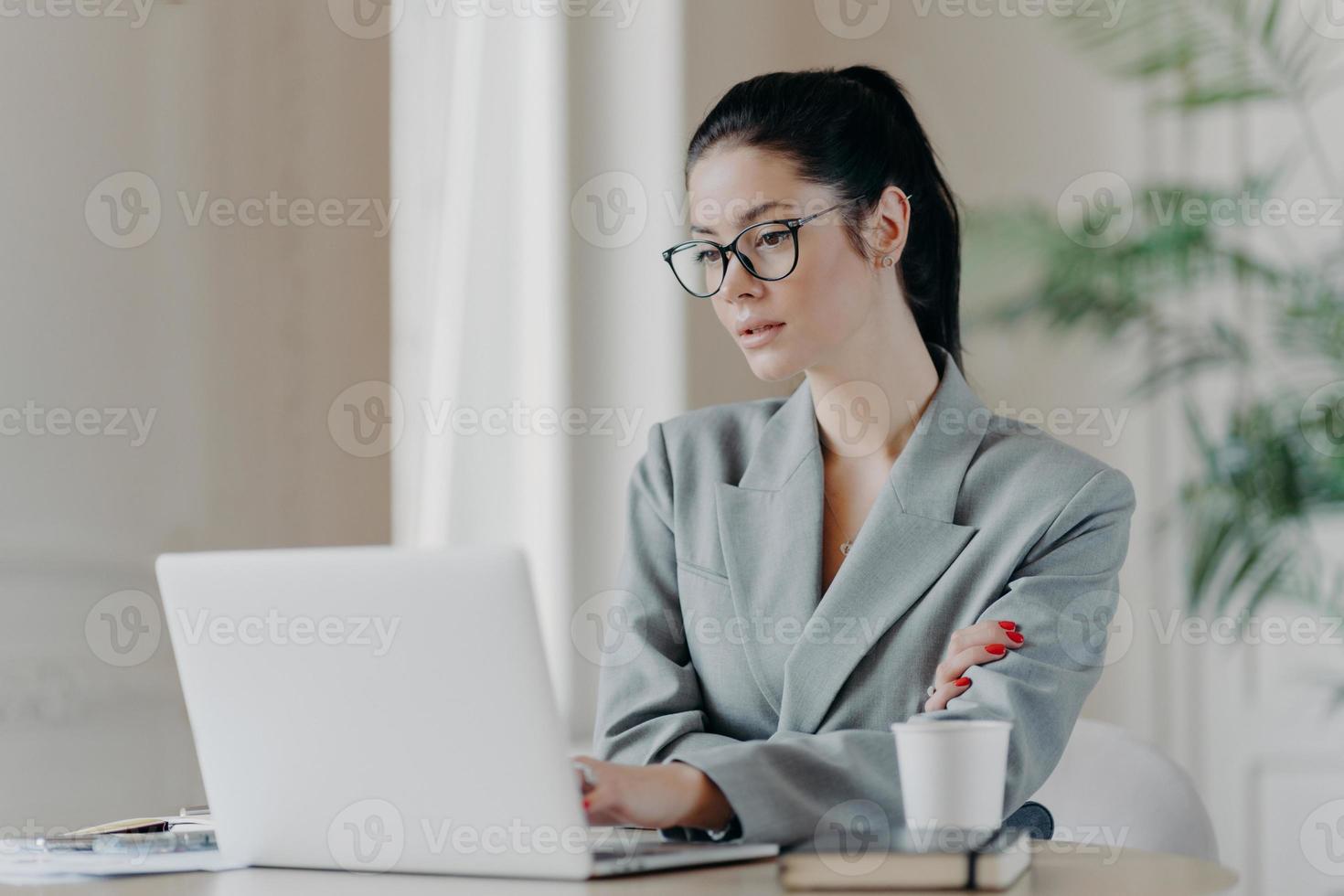 Serious female entrepreneur in eyeglasses, has dark hair combed in pony tail, works on business project, concentrated at laptop computer wears grey formal costume uses information from web resources photo
