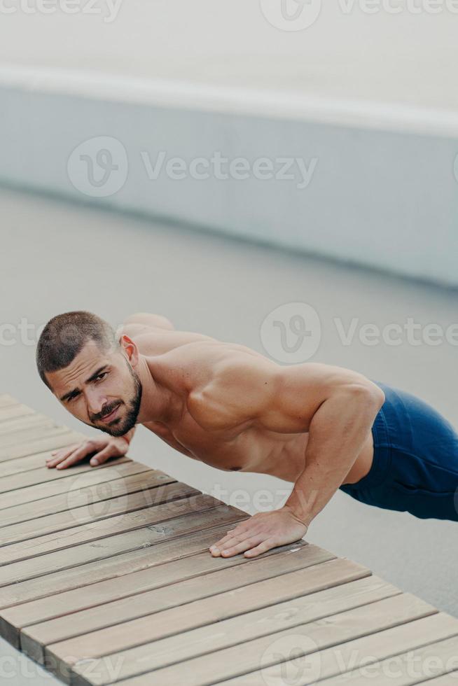 Vertical shot of muscular bearded man stands in plank pose trains chest and arms muscles has hard workout for keeping athletic body shape poses outdoor. Active lifestyle and motivation concept photo