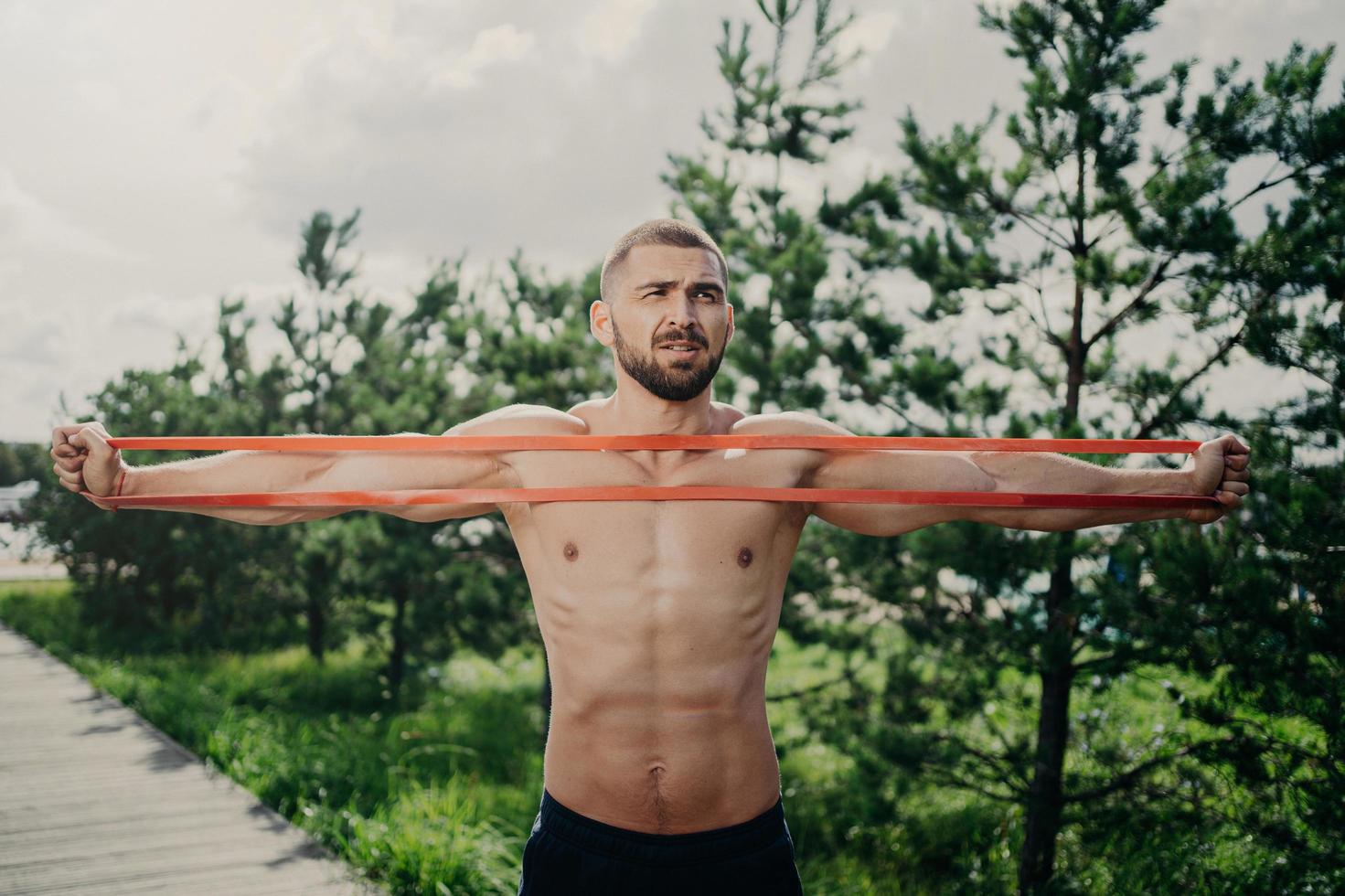 Horizontal shot of sporty man with bare chest exercises with elastic expander, gains for strong muscular body, has thick bristle, poses outside. Bodybuilder stretches band stands near green trees photo