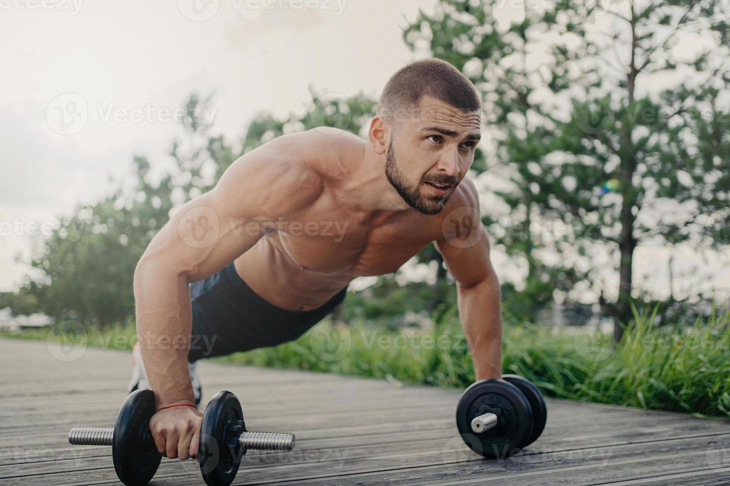Muscular male bodybuilder does push up with barbells stands in palnk pose with naked body, trains determination, goes in for sport regularly. Sport, fintess, powerlifting and bodybuilding concept photo