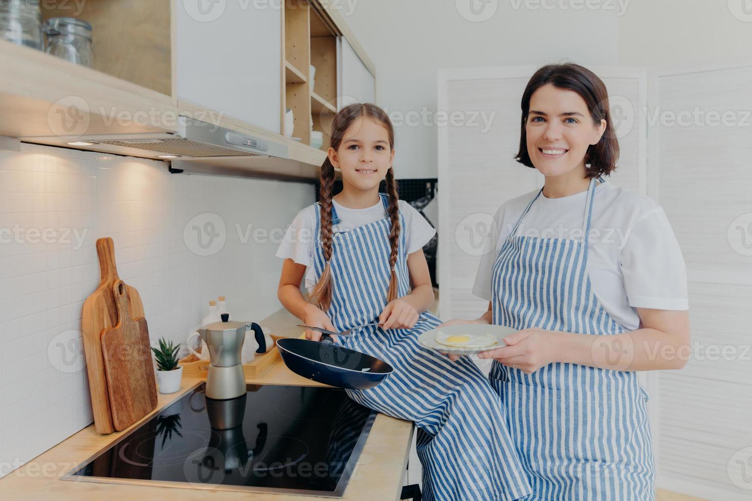 toma horizontal de madre feliz y niña pequeña miran con alegría a la cámara, ocupadas preparando huevos fritos para el desayuno o la cena, posan juntas cerca de la cocina, estando en la cocina. concepto de familia foto