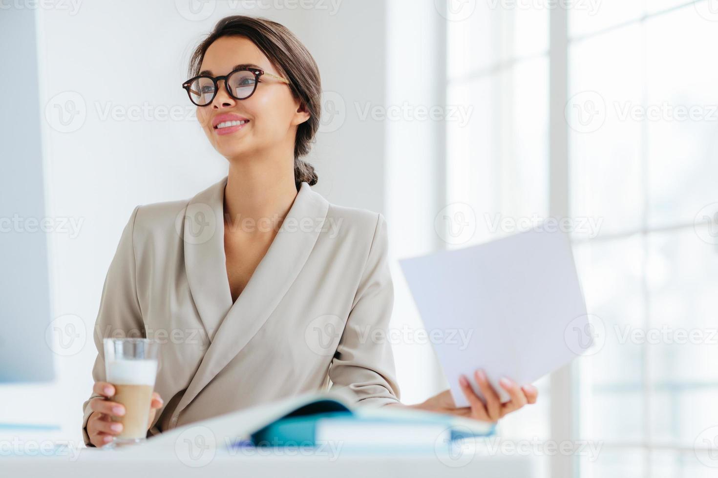 Cheerful businesswoman reads paper documents, focused aside with smile drinks milkshake sits at desktop makes business plan for project. Female executive manager in spectacles poses in coworking space photo