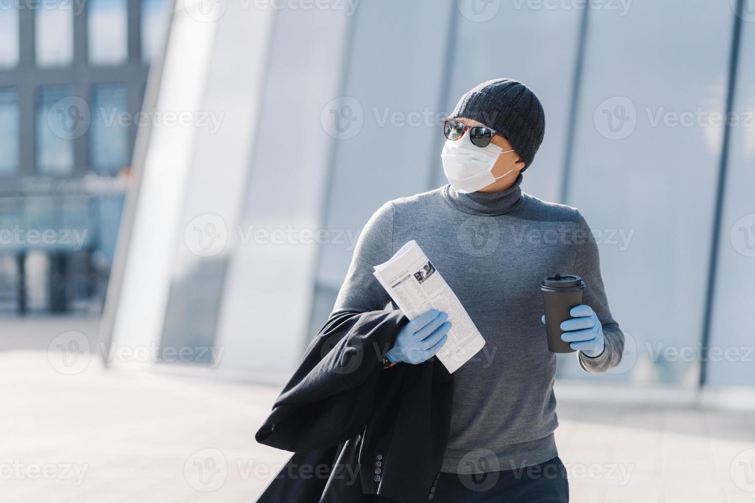 Shot of man walks outdoor, protects from flu, coronavirus spreading, drinks coffee, looks aside with pensive expression, reads newspaper, follows quarantine rules. 2019-ncov. Pandemic virus. photo