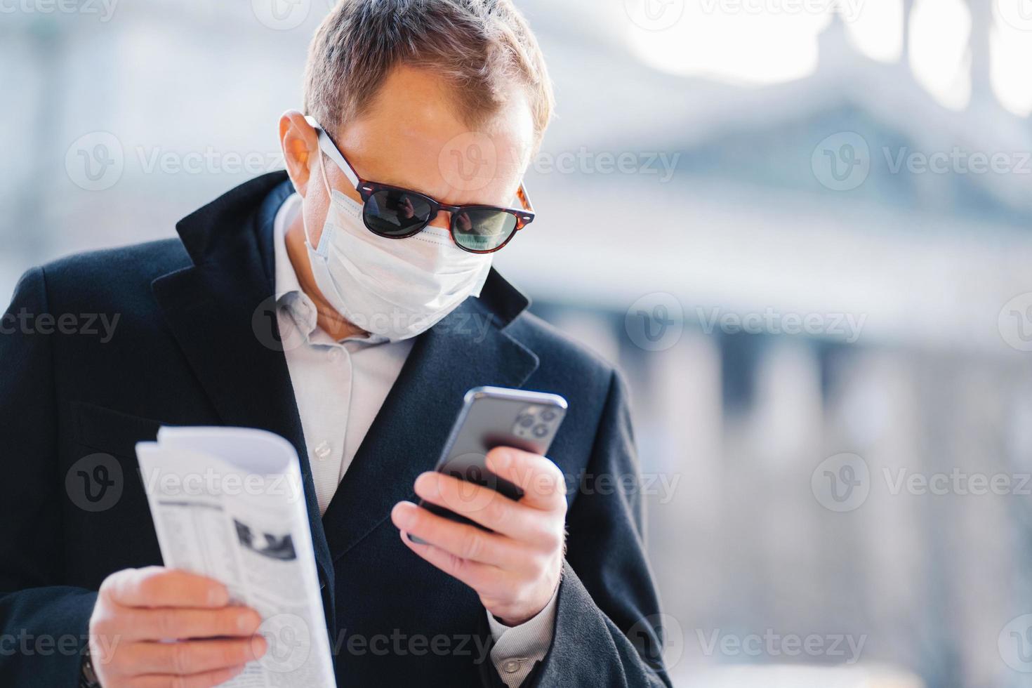 Outdoor shot of serious man manager focused in cellular display, reads news online, holds newspaper, wears medical mask to protect from coronavirus, infection with influenza virus, poses on street photo