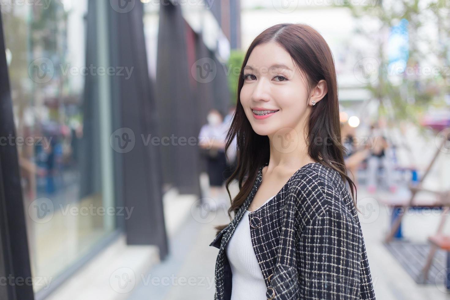 Portrait of a beautiful, long-haired Asian female in a black pattern coat with braces on teeth walking and smiling outdoors in the city. photo