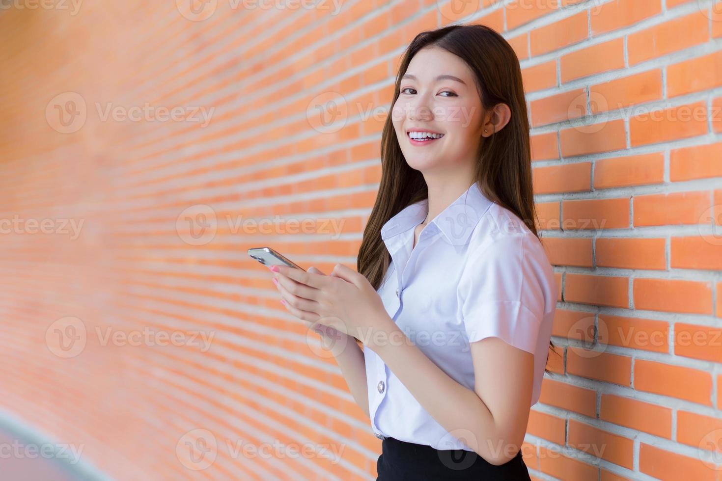 el retrato de una estudiante tailandesa asiática con uniforme sonríe alegremente mientras usa un teléfono inteligente en la universidad con paredes de ladrillo como fondo. foto