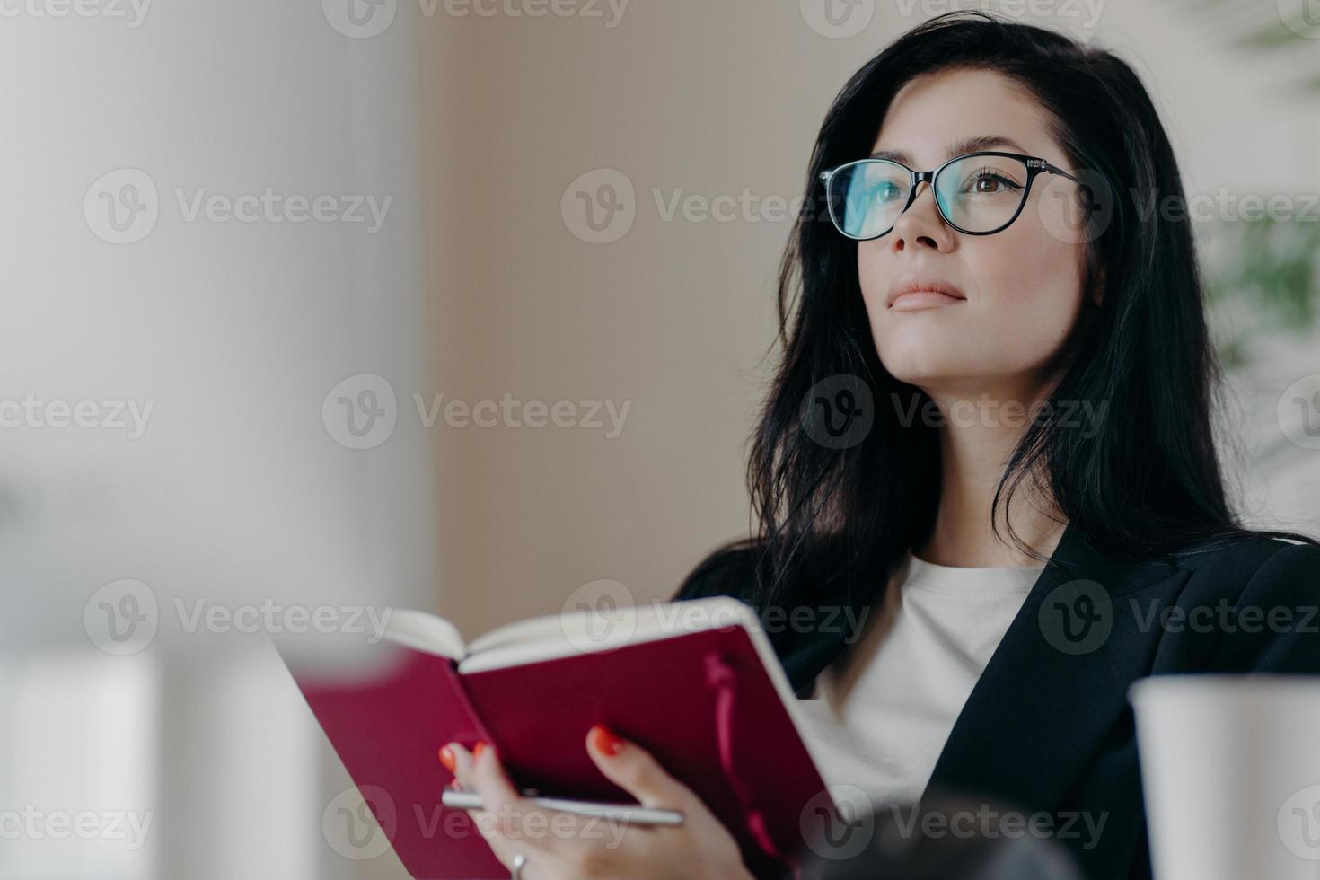 Beautiful woman with thoughtful expression, holds opened notepad, writes time management, makes plans for productive day, wears eyeglasses, poses indoor. Self employed female writes down ideas photo