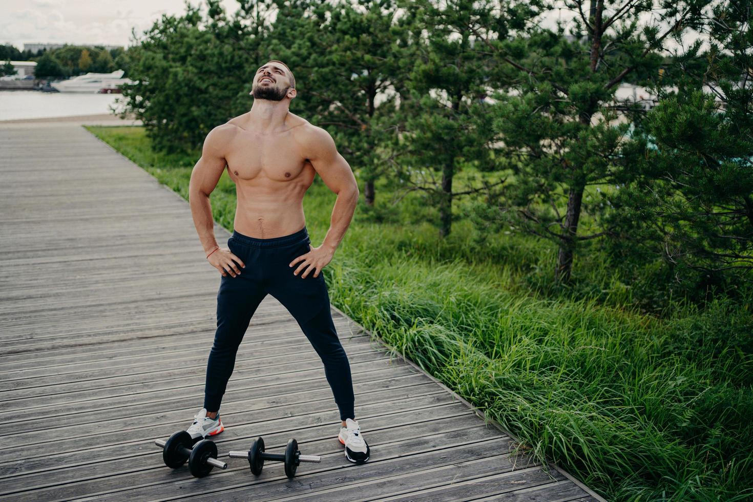 Bearded muscular man keeps hands on waist, raises head and breathes fresh air, takes break after doing workout, trains with barbells. Professional weightlifter works out in nature. Exercising concept photo