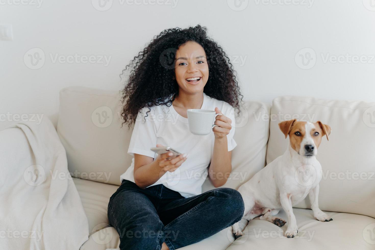 Delighted dark skinned woman poses in modern apartment, sits on comfortable sofa with pet, drinks coffee, uses mobile phone for online communication, being in good mood, flipping news, uses app photo