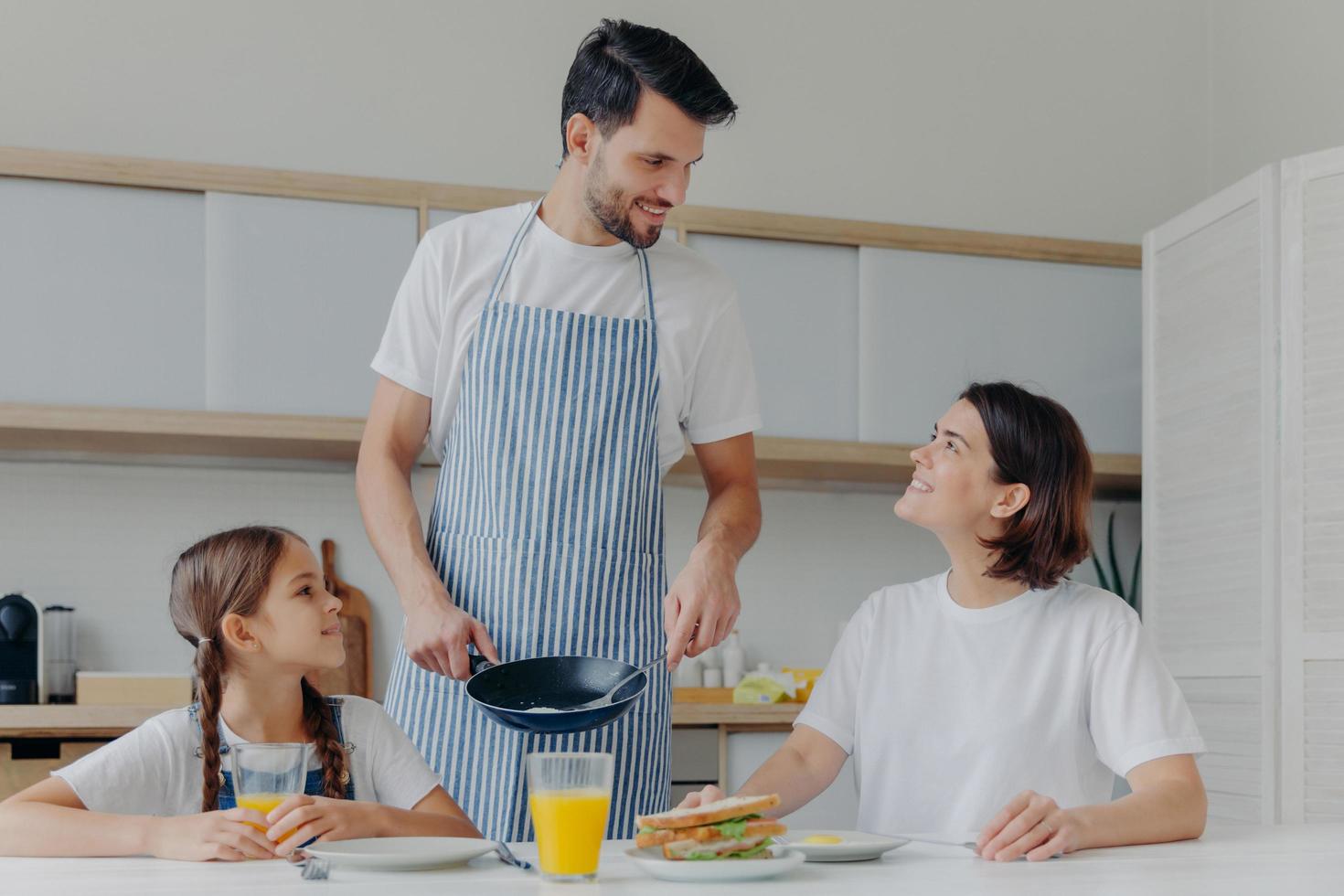 feliz padre, madre e hija se reúnen en la cocina, comen un delicioso desayuno, papá preparó huevos fritos, de buen humor, listos para comenzar un nuevo día. encantadora familia disfruta de una sabrosa comida en casa foto