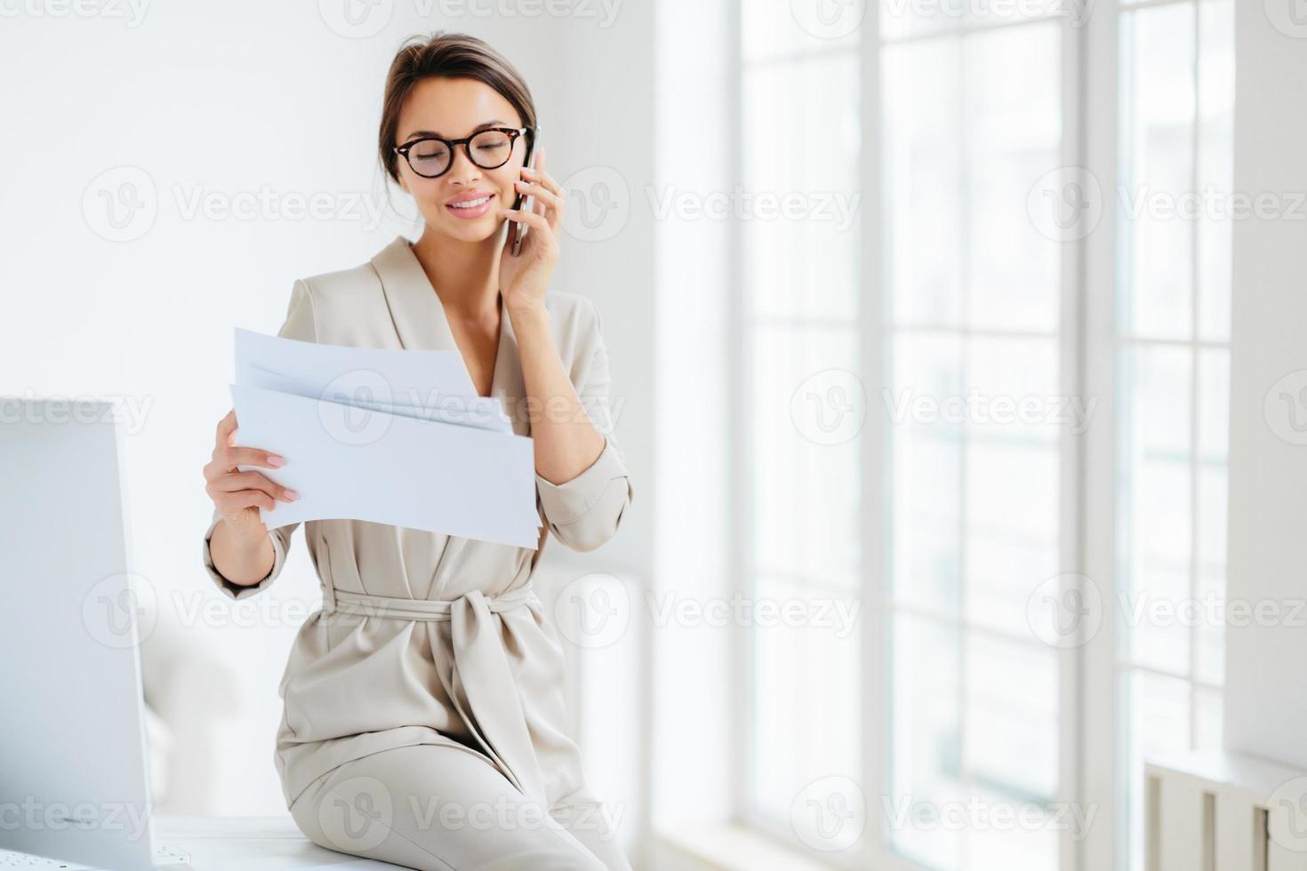 Attractive businesswoman reads papers or business documents, has telephone talk with business partner, examines paperwork before meeting, sits at desktop, smiles happily, works with documentation photo
