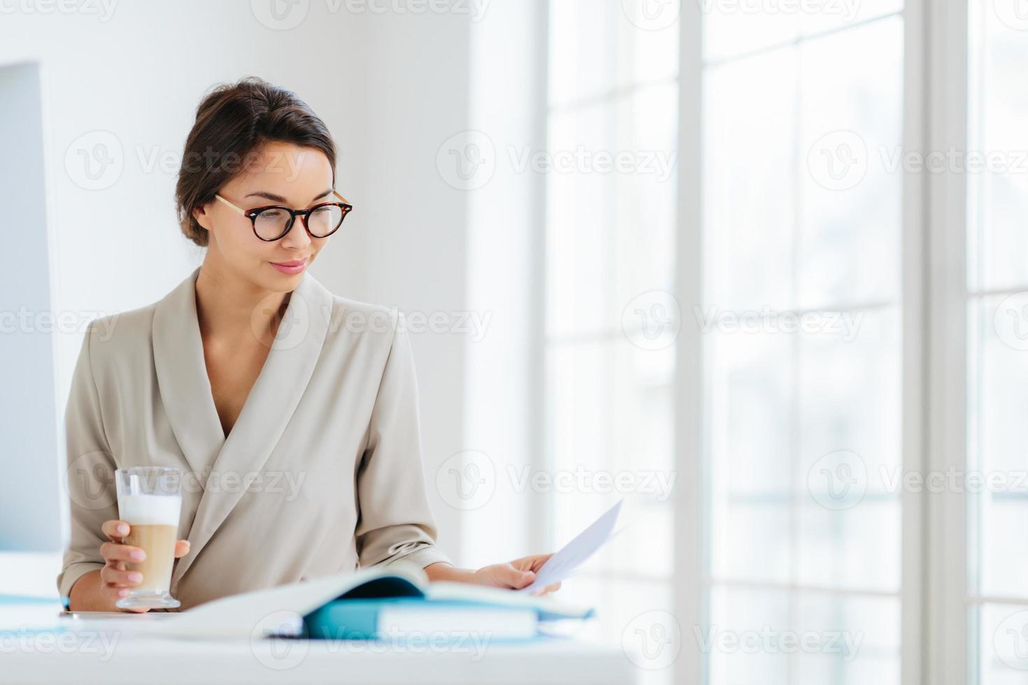 Woman economist checks information on business paper, wears spectacles and elegant suit, drinks milkshake, poses against office interior, being in cabinet, prepares startup project. Working process photo
