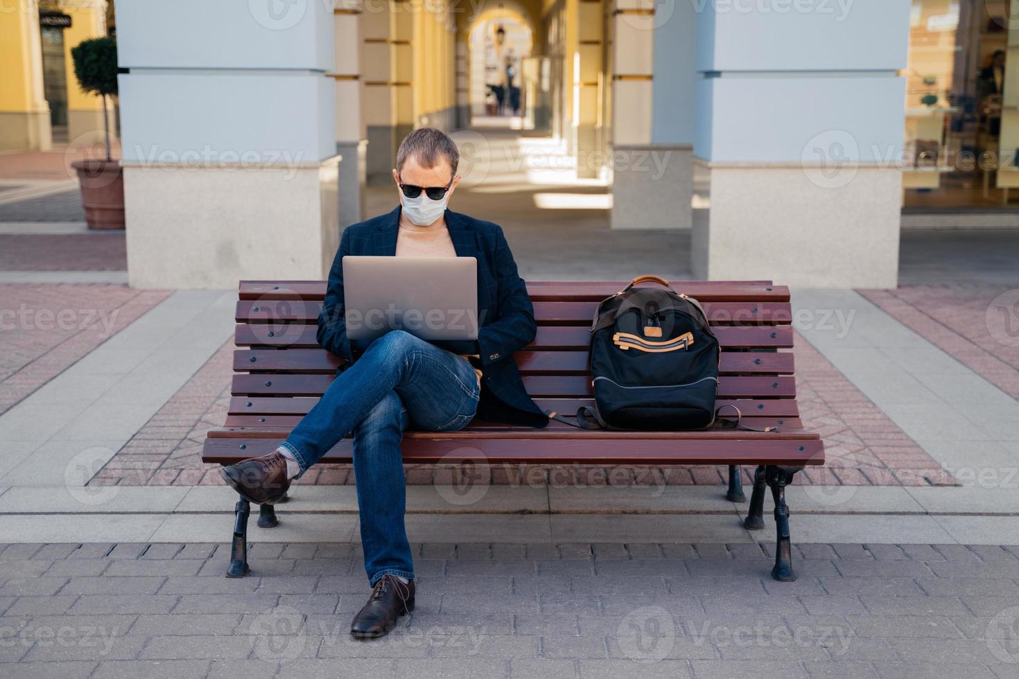 Photo of busy man freelancer in mask does distance work, keyborads on laptop computer, sits at wooden bench outside, avoids crowded places during quarantine time. Traveling and coronavirus concept