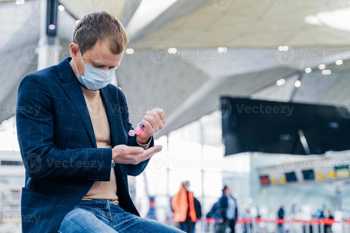 Photo of man in medical face mask, uses disinnfectant spray for hands, protects from coronavirus in crowded place, poses in airport, waits for flight. Health care concept. Sanitation, virus prevention