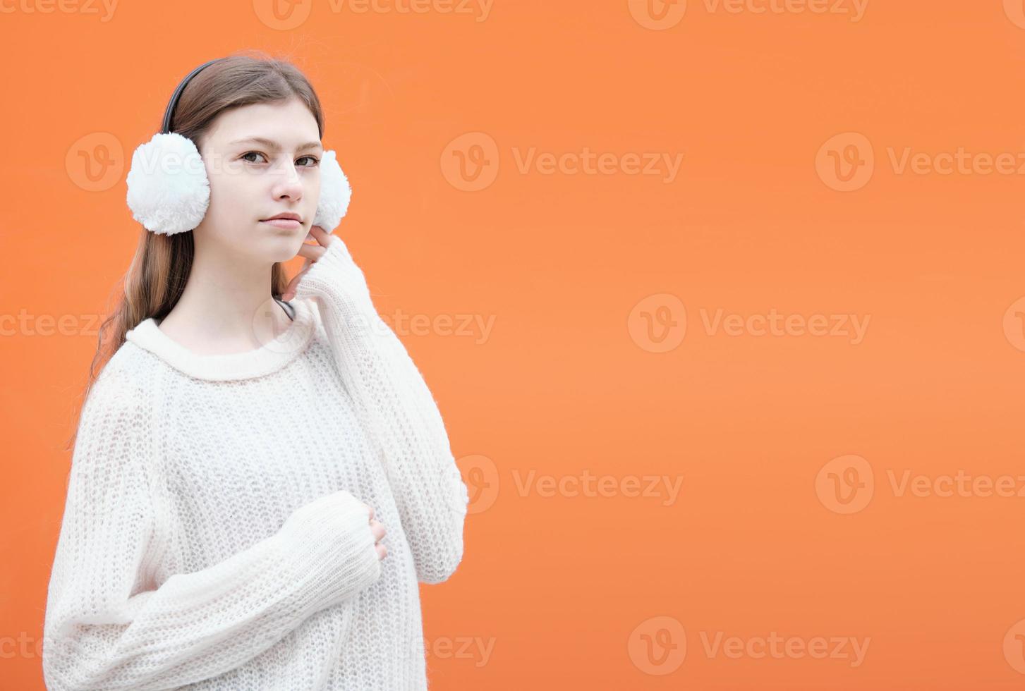 chica adolescente de moda con orejeras blancas con suéter y de pie contra un fondo naranja. foto