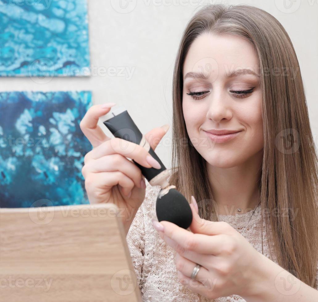 mujer joven aplicando base de maquillaje en una esponja en casa. automaquillaje, gente real. la mujer se prepara para salir. tono de piel perfecto foto