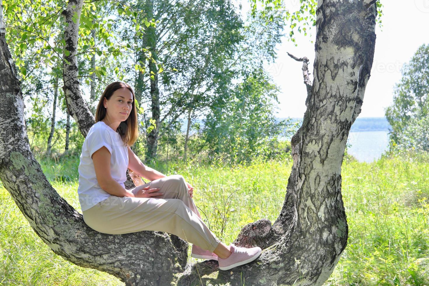 beautiful slim woman getting rest on a tree in a forest. mid adult woman wearing casual clothes sitting alone in a forest in summer. enjoying active lifestyle photo