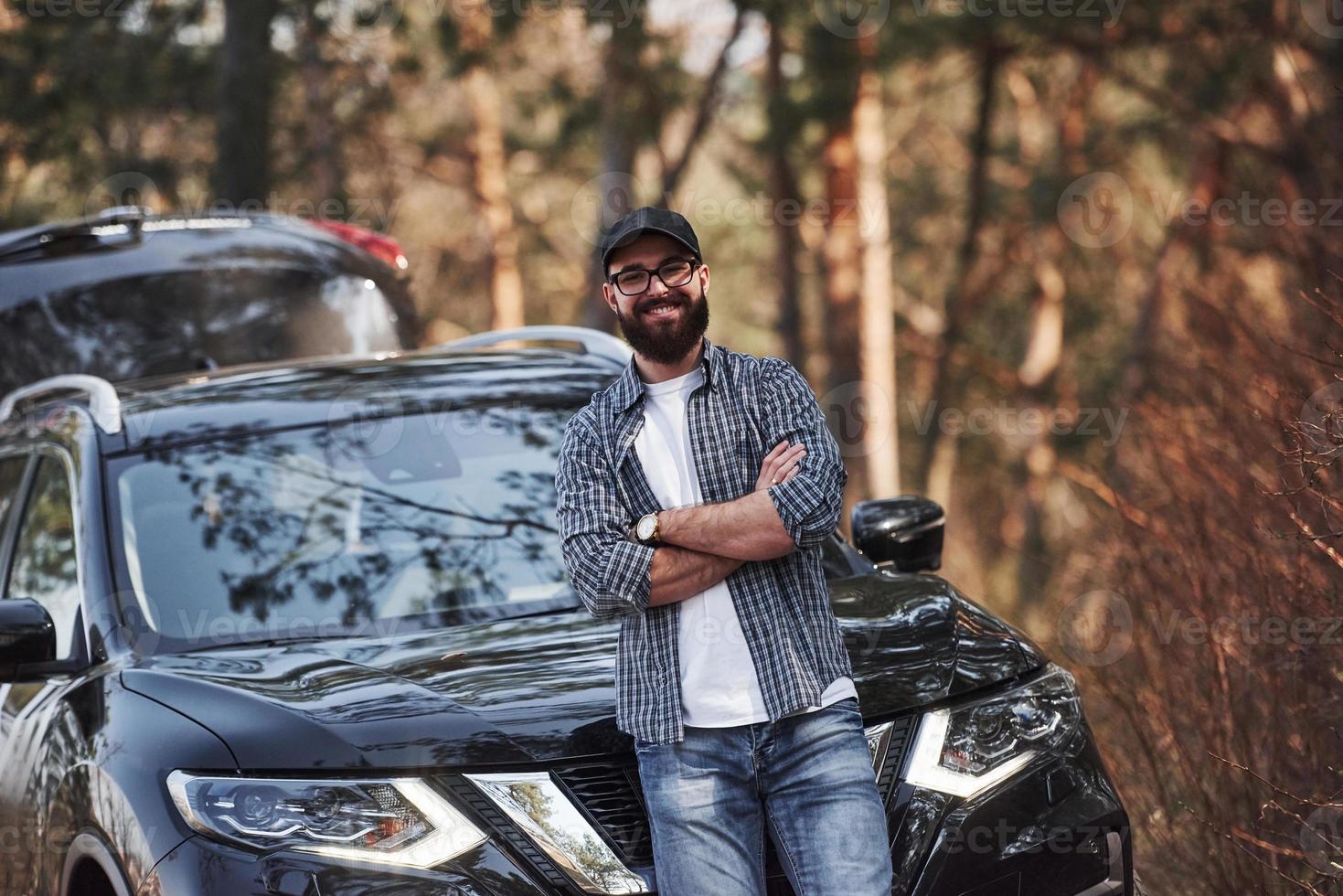 en anteojos negros. hombre barbudo cerca de su nuevo auto negro en el bosque. concepto de vacaciones foto