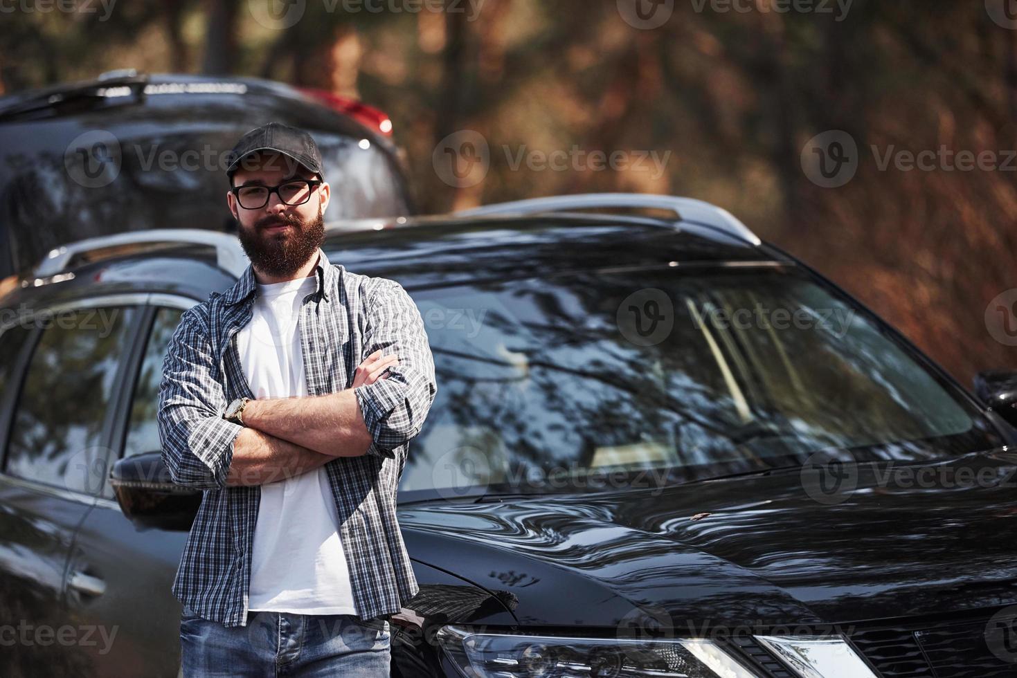 con los brazos cruzados. hombre barbudo cerca de su nuevo auto negro en el bosque. concepto de vacaciones foto