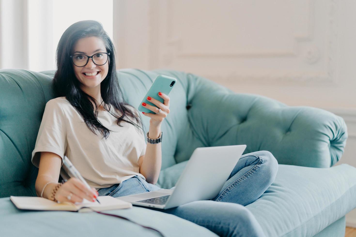 Pleased young female designer with dark hair creats new project, holds modern smartphone in hands, makes notes in notepad, poses on sofa, uses laptop computer connected to wireless internet. photo