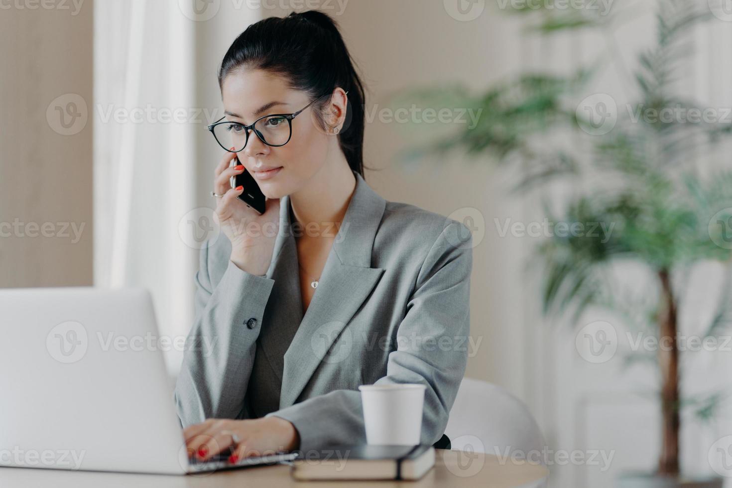 Serious brunette woman keyboards information, concentrated into laptop computer, dressed in formal outfit, poses at coworking space, works remotely, involved in working process during daytime. photo