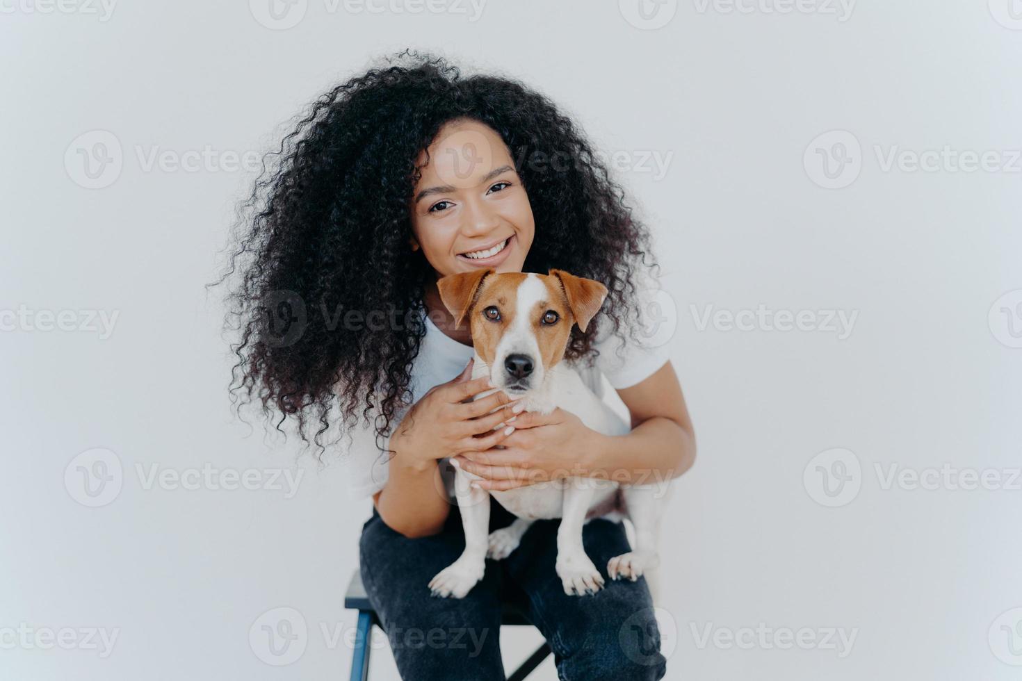 una mujer afroamericana sonriente posa con un animal doméstico, mira alegremente a la cámara, abraza a un perro, se sienta en una silla, aislada sobre fondo blanco. dos miembros de la familia. relax hogareño. juntos para siempre foto