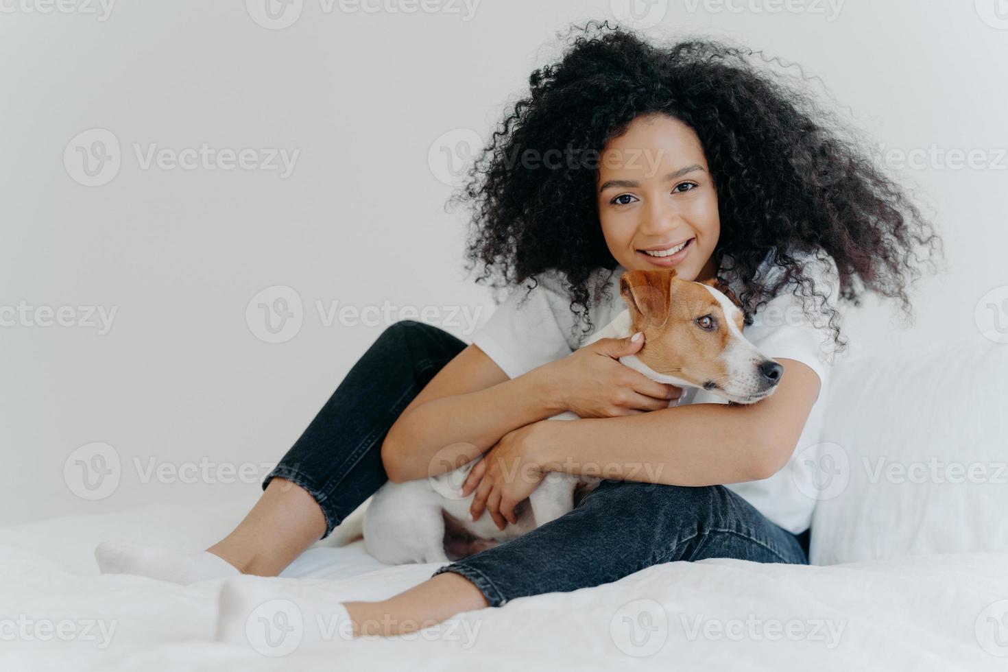 Photo of lovely young female pet owner poses in clean white bedroom, hugs dog, plays with best friend, wears casual clothing, has cheerful expression. People, animals, love, friendship concept