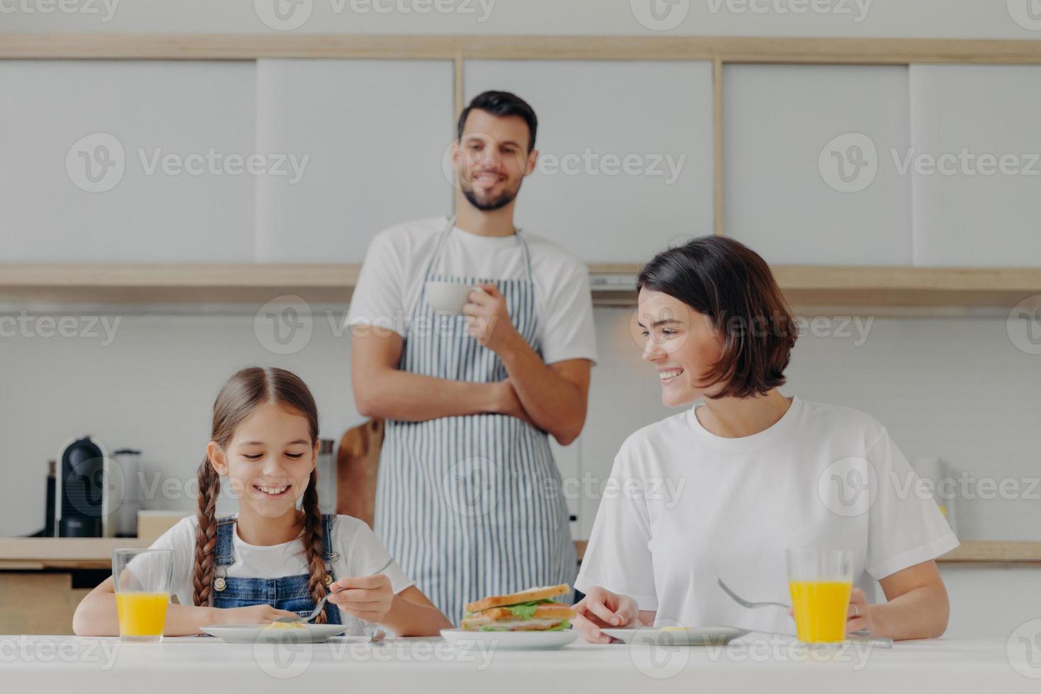 Happy mother talks to daughter while have breakfast. Father stands behind, prepared delicious dish for family. Friendly family members meet at kitchen during weekend, enjoy nice conversation photo