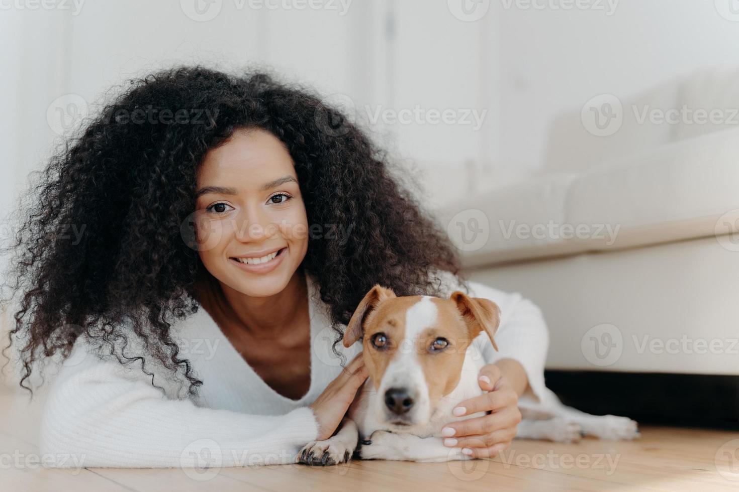 Pretty girl with Afro hair, lies on floor with dog, expresses pleasant emotions, poses in living room near couch, bought pet in new apartment. Woman host with beloved animal at home, share good moment photo