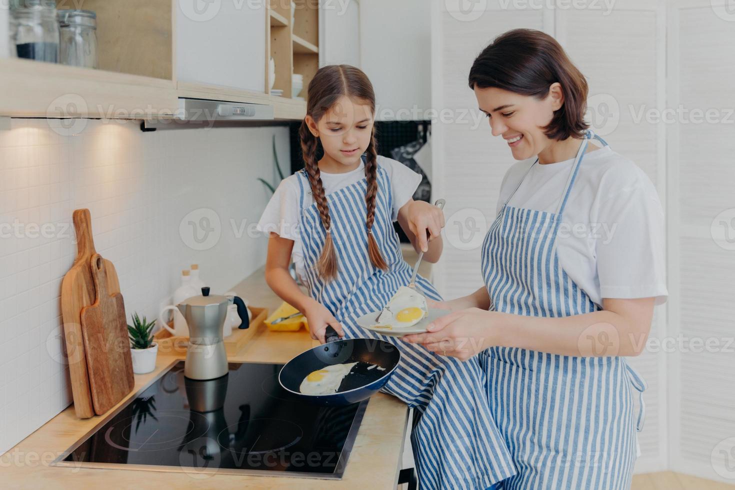 foto de una madre alegre y ocupada y su hija posan cerca de la estufa, sirven el desayuno para la familia, fríen huevos en una sartén, usan delantales, posan en la cocina doméstica. la madre le enseña al niño cómo preparar la comida fácilmente