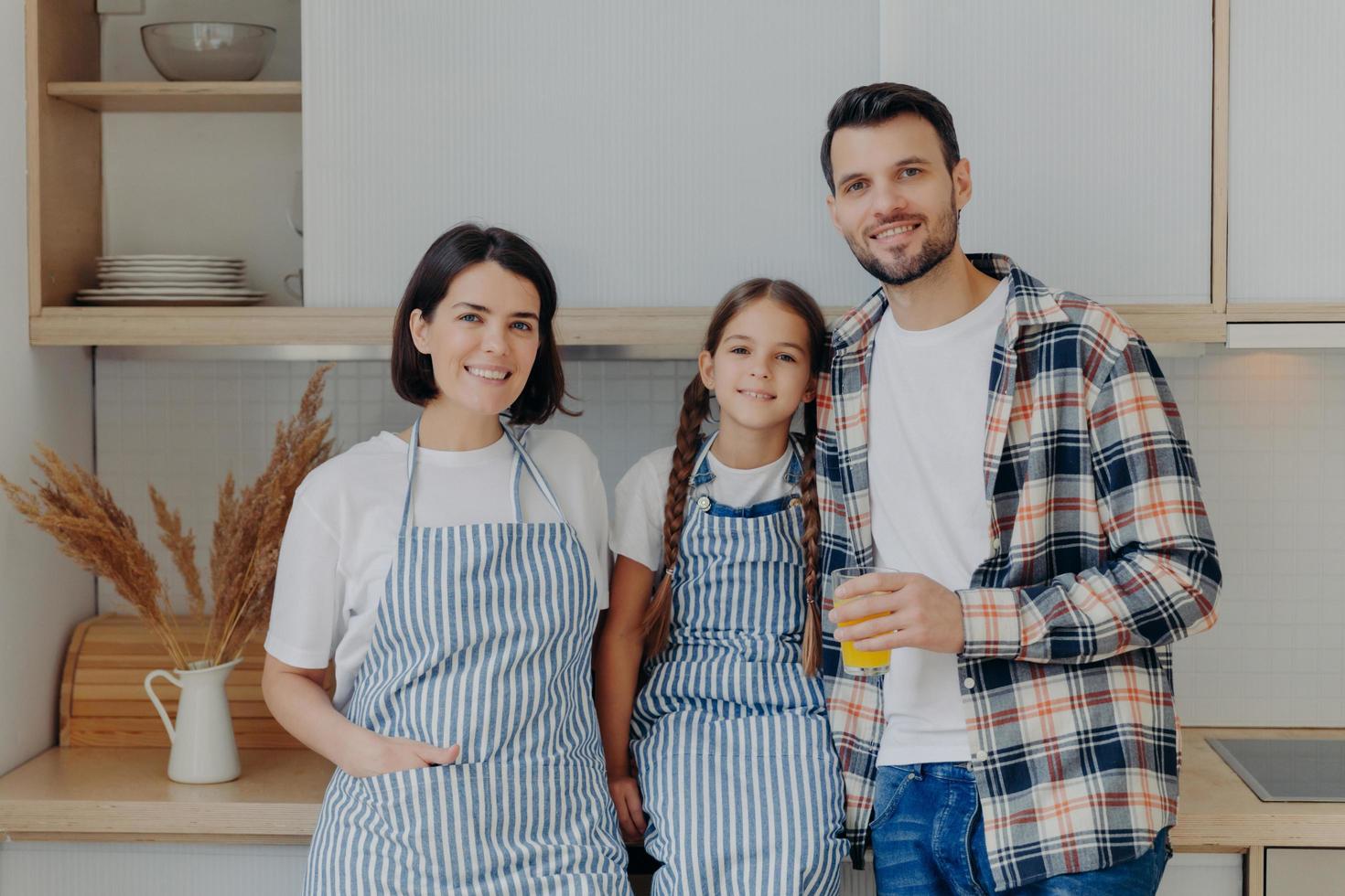 Happy family have great time together, pose in modern kitchen at home. Glad man in checkered shirt holds glass of juice, small child with pigtails, pretty housewife in apron. Girl with parents photo