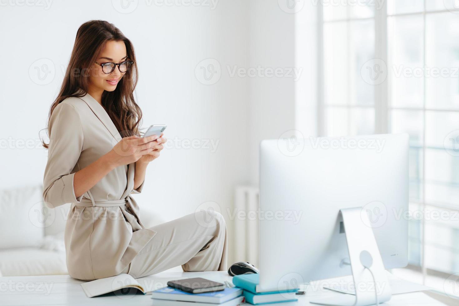 Elegant woman in beige formal suit reads news on website attentively, sits at desktop alone in cabinet, computer monitor and notepads around. Female employee uses modern smartphone in office photo
