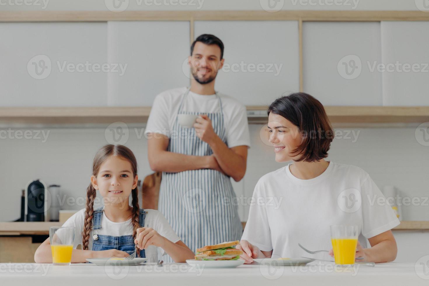 un niño pequeño encantador y su madre desayunan juntos, se sientan en la mesa de la cocina, comen una comida deliciosa, el padre se para en el fondo, usa delantal y toma café. la familia amistosa se reúne en la cocina foto