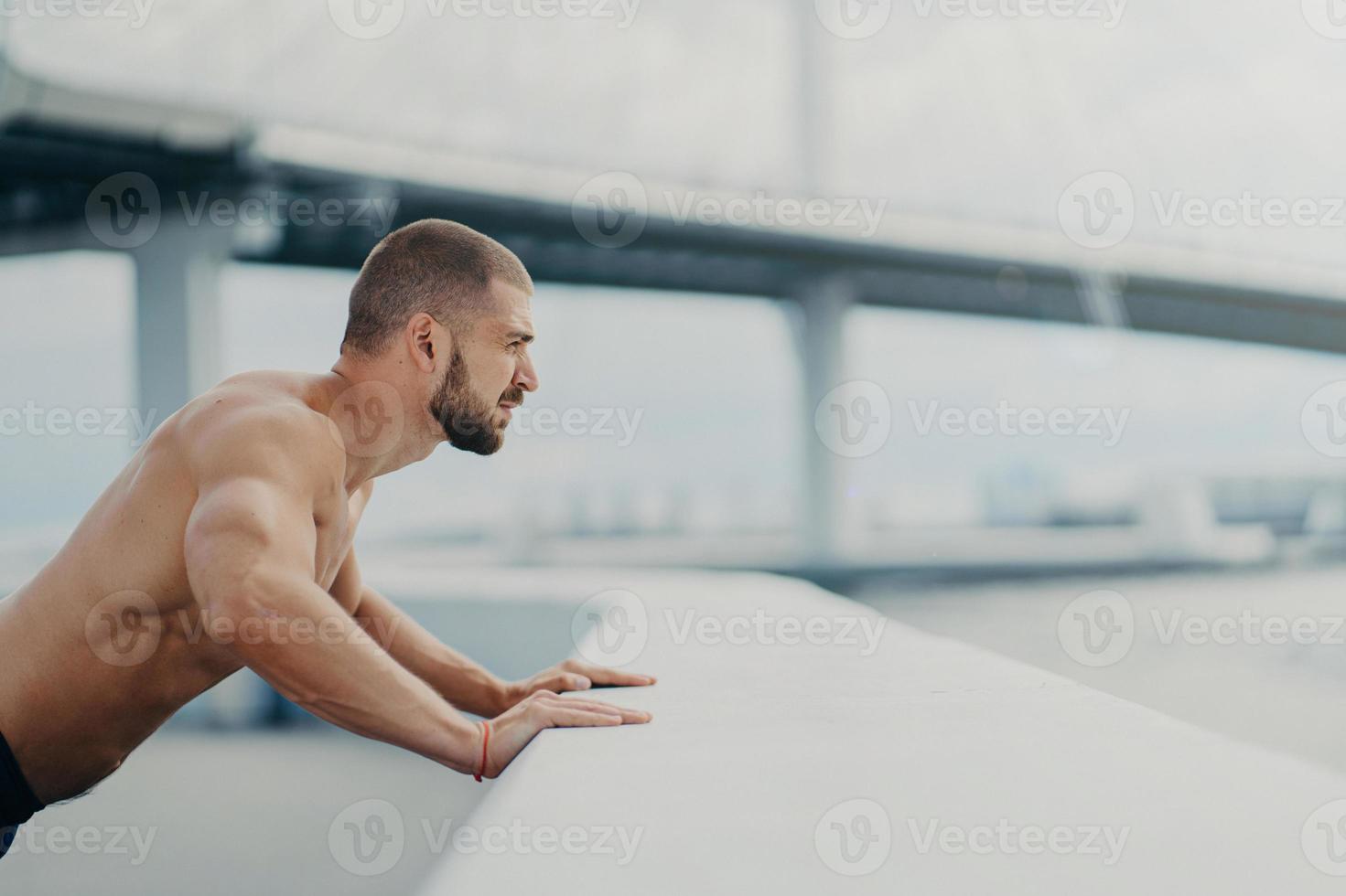 Sideways shot of handsome muscular man with thick beard does push ups exercise has workout outdoor leans on bridge fence engaged in physical activity poses with bare torso has strong muscles photo