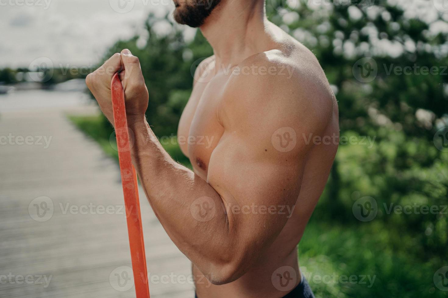 Cropped shot of unrecognizable muscular man has workout biceps exercise stands in profile shows strong muscles, uses resistance band, strives for strong body. Bodybuilding, motivation concept photo