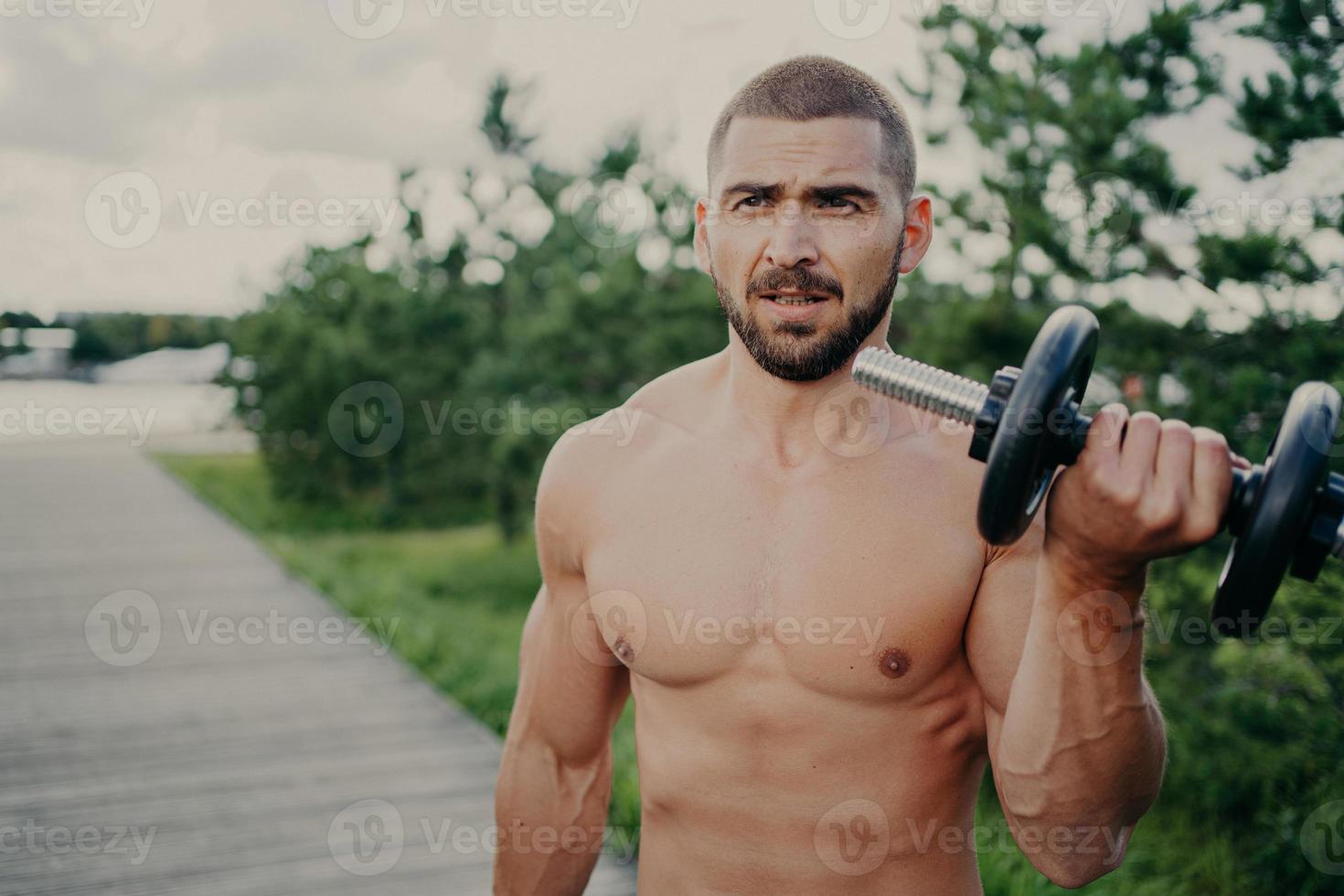 Horizontal shot of handsome muscular man lifts barbell outdoor, has athletic torso, gets ready for weight lifting training, has muscular arms, poses against street background. Developing strength photo