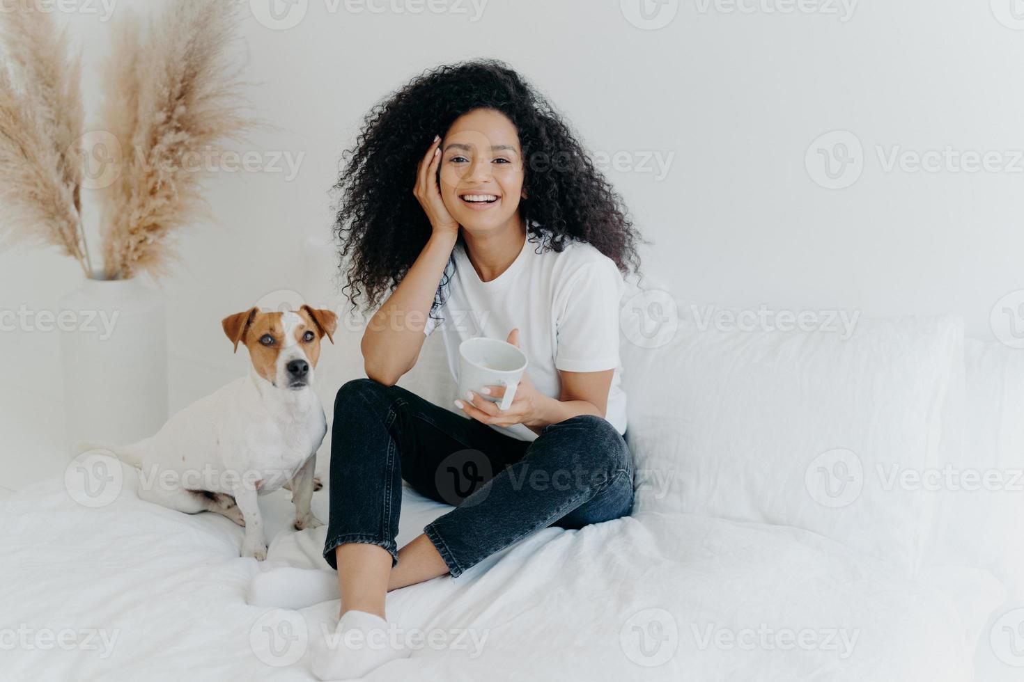 Happy attractive ethnic woman with curly hair wears white t shirt, jeans and socks, smiles pleasantly, drinks tea in comfortable bed, poses with dog, have lazy weekend. People, rest, animals concept photo