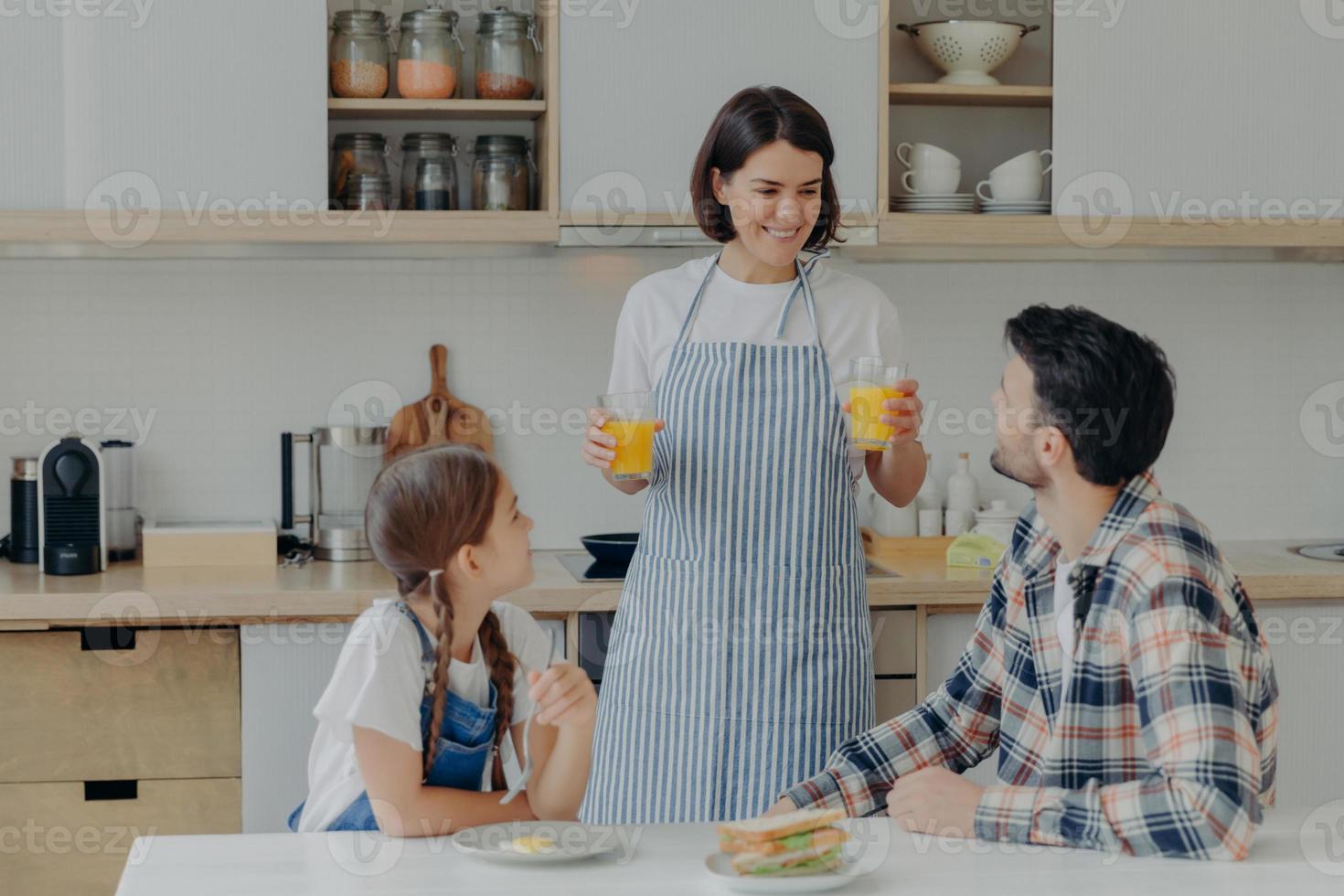 Lovely happy mother wears apron, holds two glasses of orange juice, talks to husband and daugher, prepares breakfast. Three membered family have dinner time at kitchen. Togetherness concept. photo