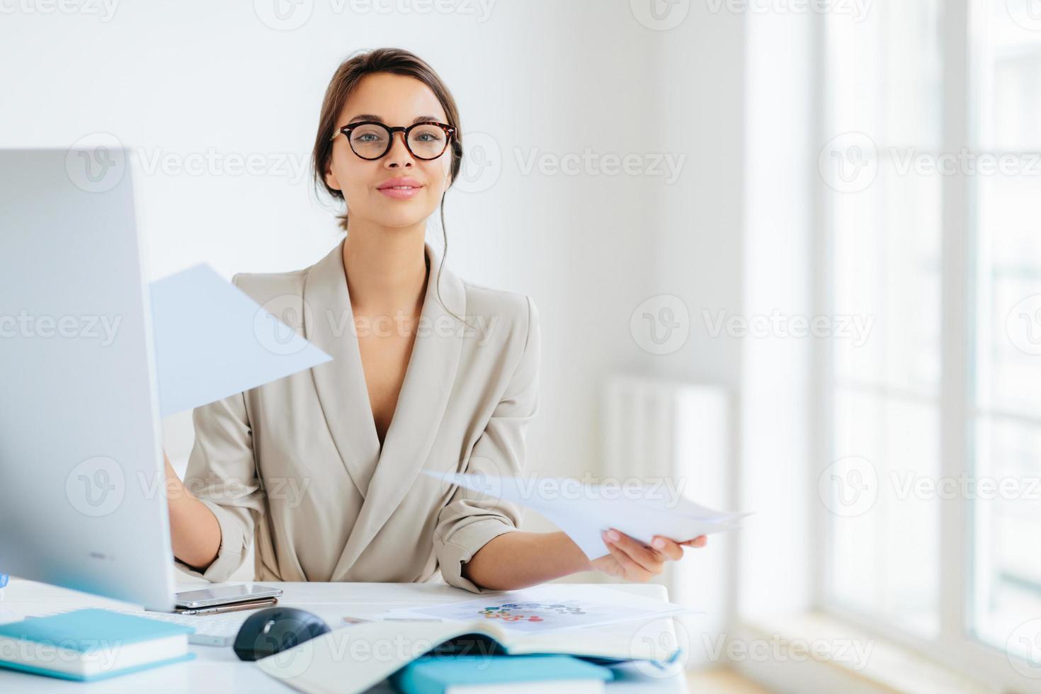 Busy successful female manager works with papers in office, poses at desktop, wears spectacles and formal outfit, busy preparing report, looks confidently at camera. Businesswoman reviews documents photo