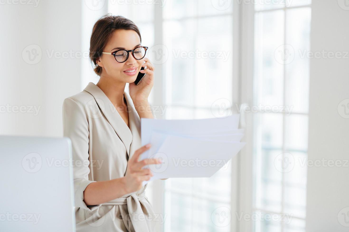 Positive woman in formal clothes, calls to business partner, discusses financial startup project, reads information from papers, wears optical glasses, stands in office, works at international company photo