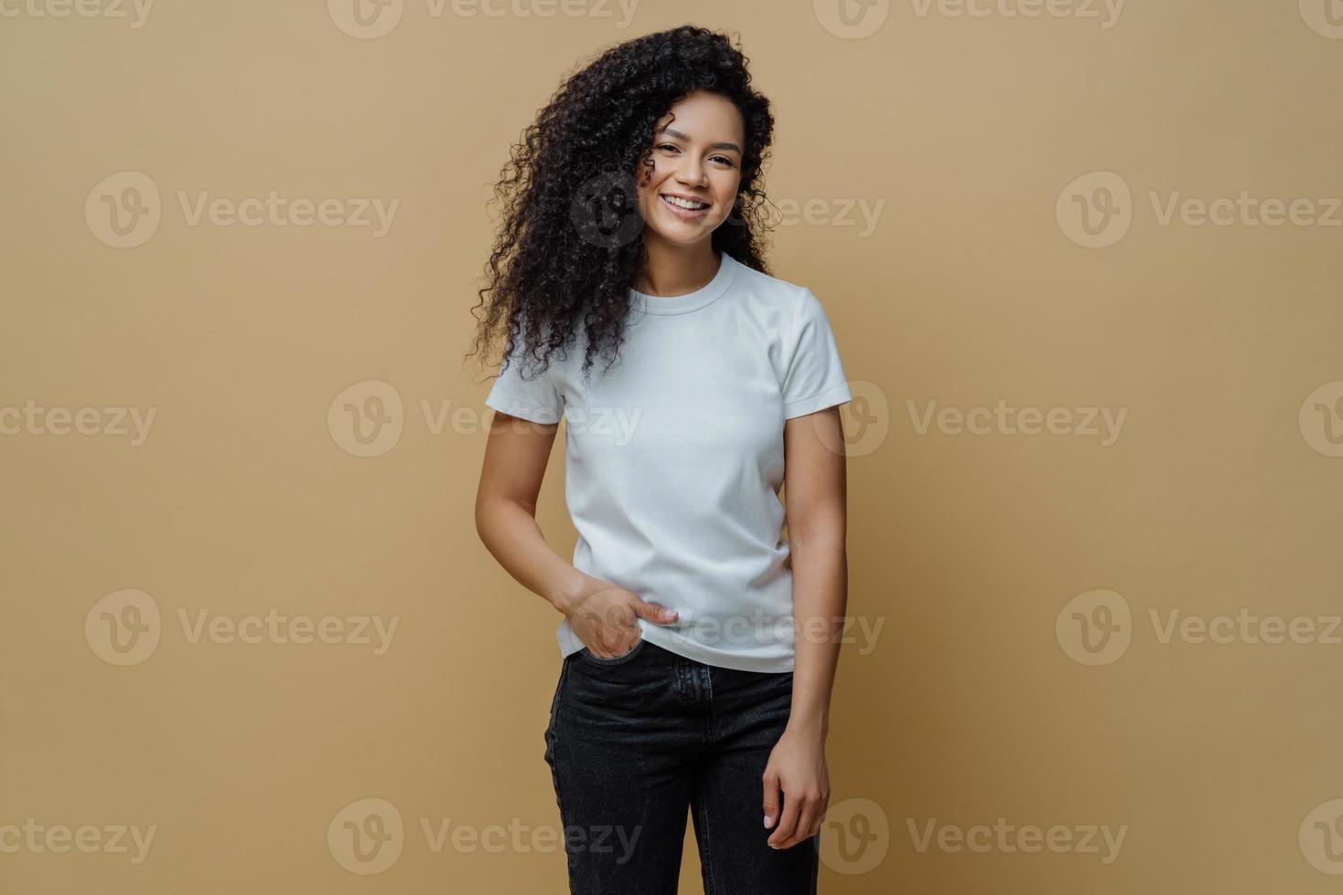 Photo of slim cheerful African American woman with curly hair, smiles happily, being in good mood, wears white t shirt and jeans, keeps hand in pocket, has slim figure, isolated on beige background