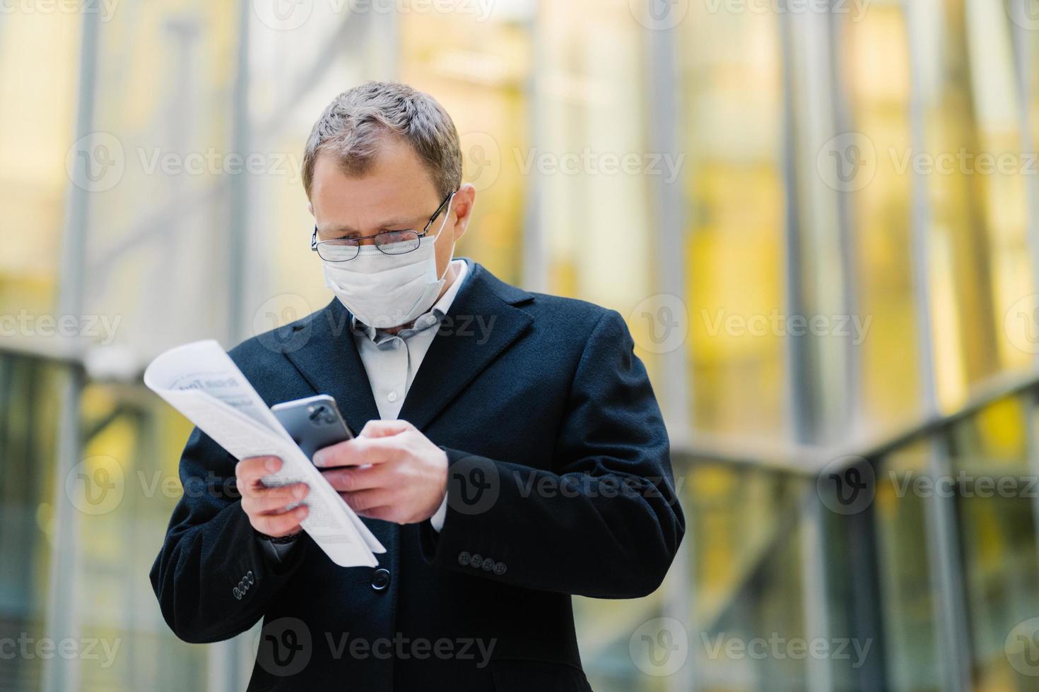 Coronavirus pandemic concept. Photo of male manager focused in smartphone device, sends text messages, wears spectacles and medical mask, holds papers, walks at street during quarantine, pandemic