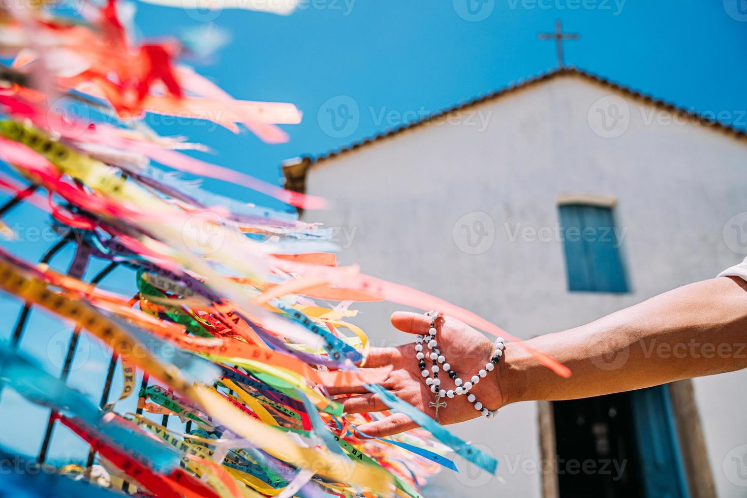 la mano de un hombre haciendo un pedido con cintas brasileñas en la cerca de una iglesia en arraial d'ajuda, bahia, brasil. centrarse en cintas de colores foto