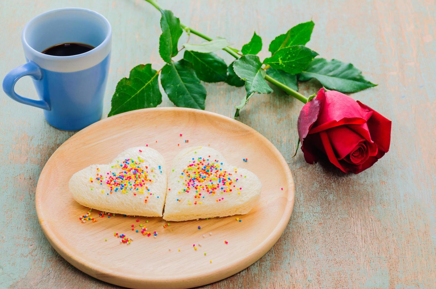 Couple of heart shape white bread on wooden plate with coffee cup and red rose. Photo is focused at the bread.