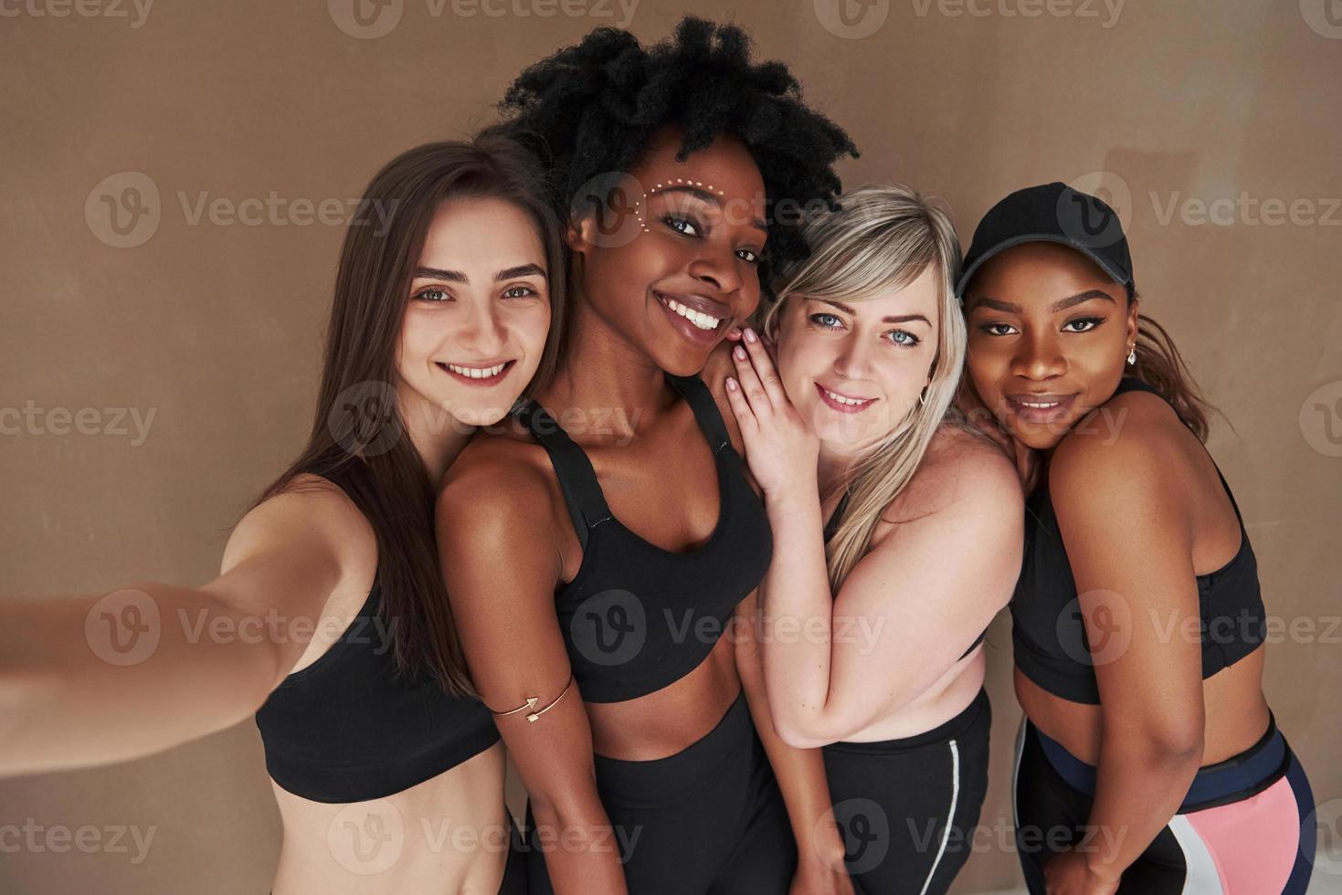 Holding camera on elongated hand. Group of multi ethnic women standing in the studio against brown background photo