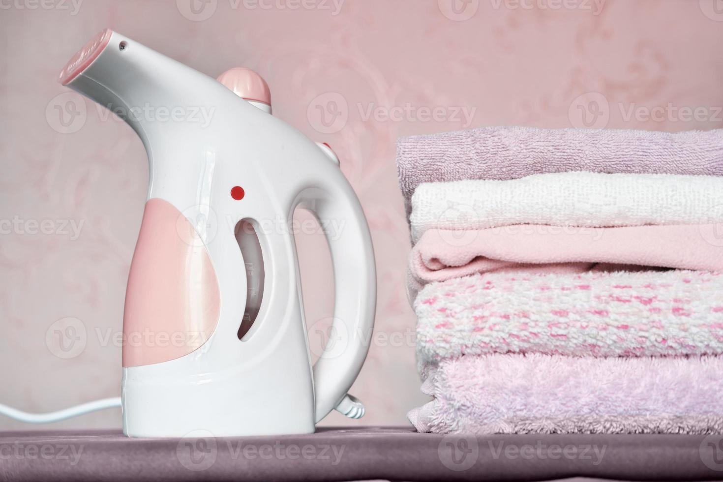 vaporizador de ropa eléctrico portátil y montón de toallas dobladas en la tabla de planchar. limpieza y hogar, concepto de tareas. foto